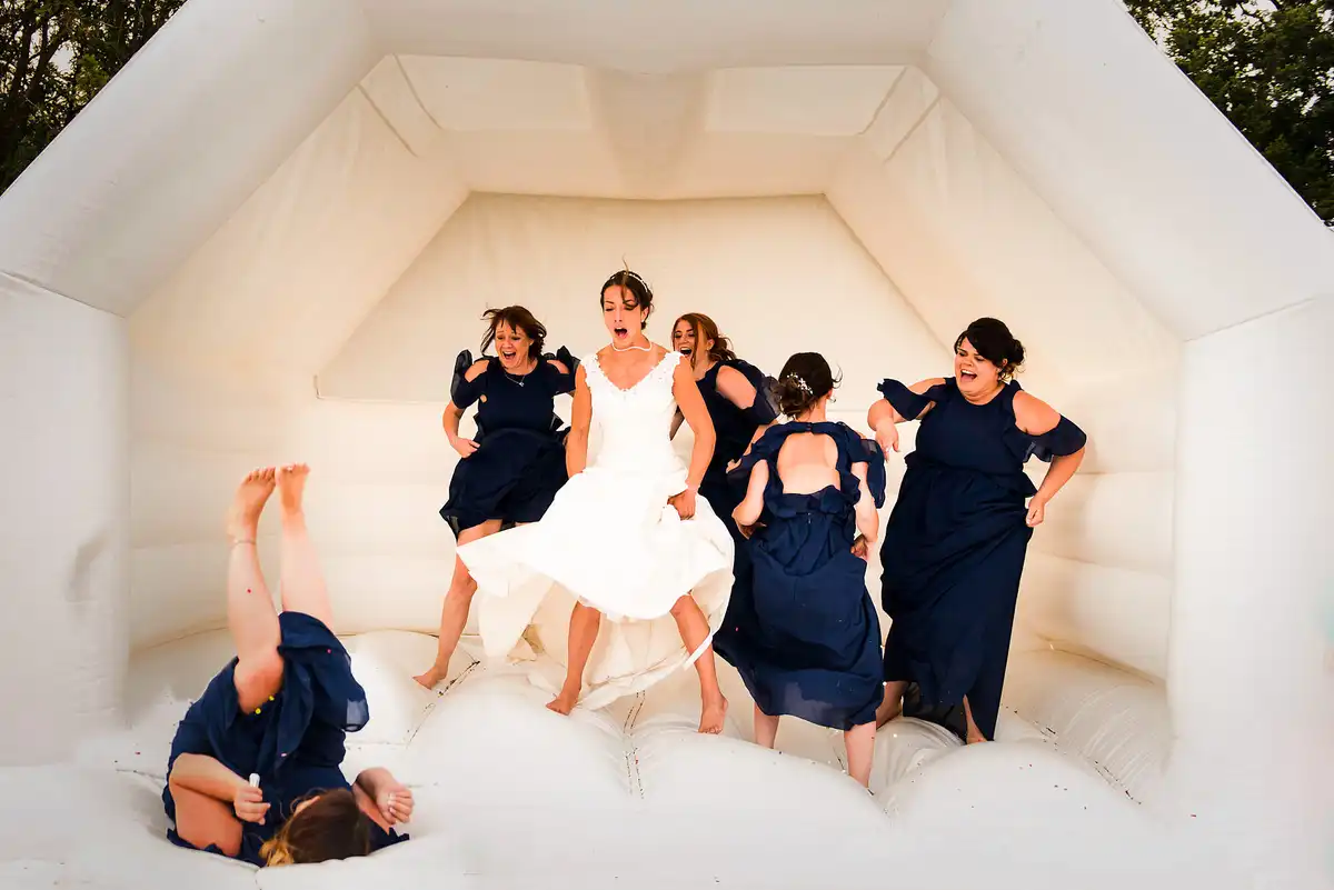 Bride and bridesmaids in navy bounce inside a white bouncy castle, one falling with her legs in the air.