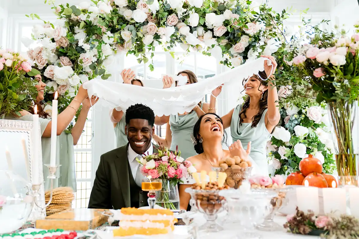 Couple laughing at a flower-covered table as bridesmaids in sage hold a white cloth over their heads.