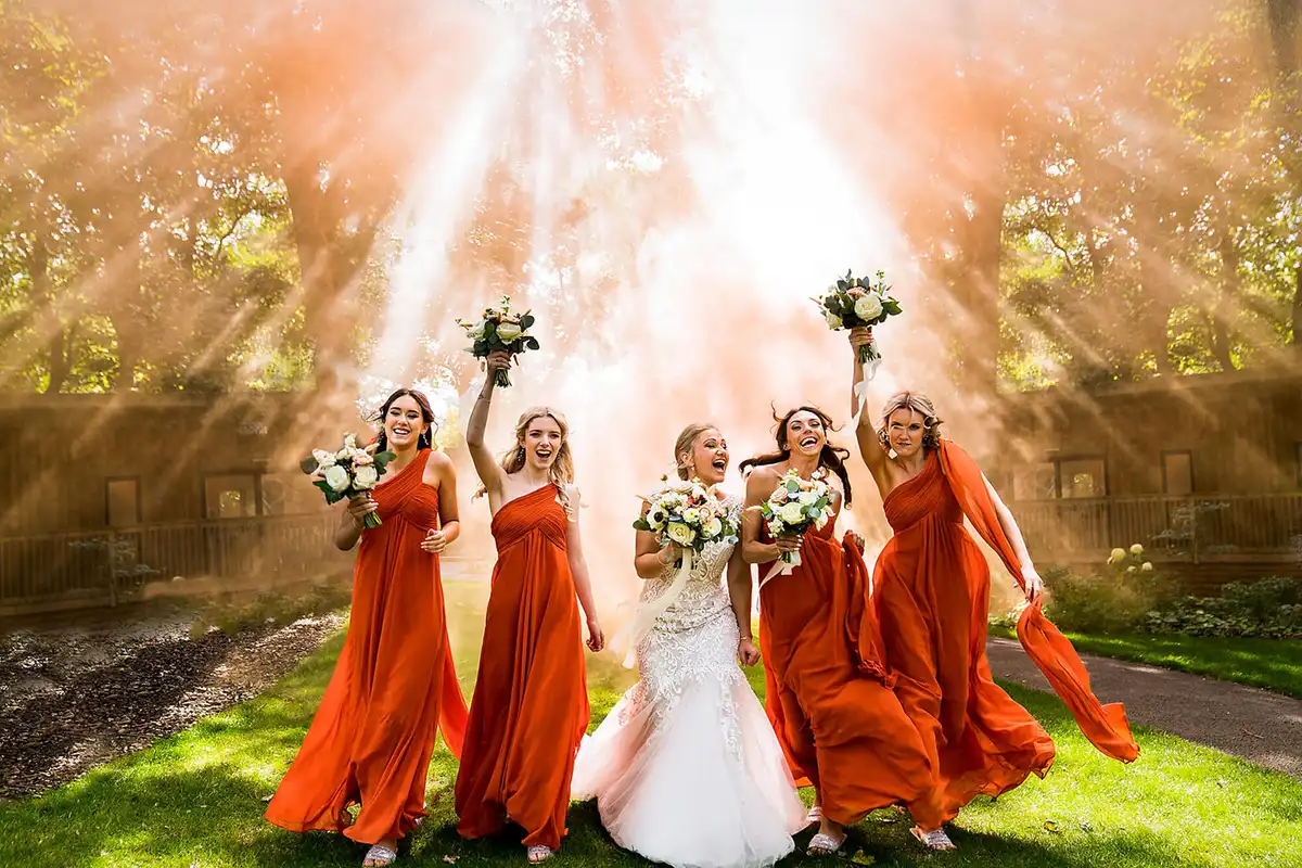 Bride and four bridesmaids in burnt orange laugh with bouquets raised in orange smoke and sunbeams.