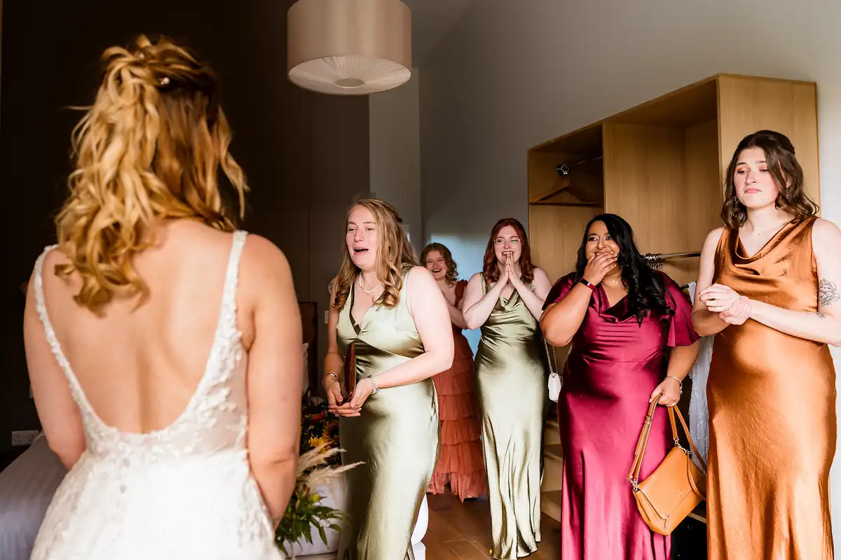 Bridesmaids in satin gasp and tear up as they see the bride, who stands with her back to the camera.
