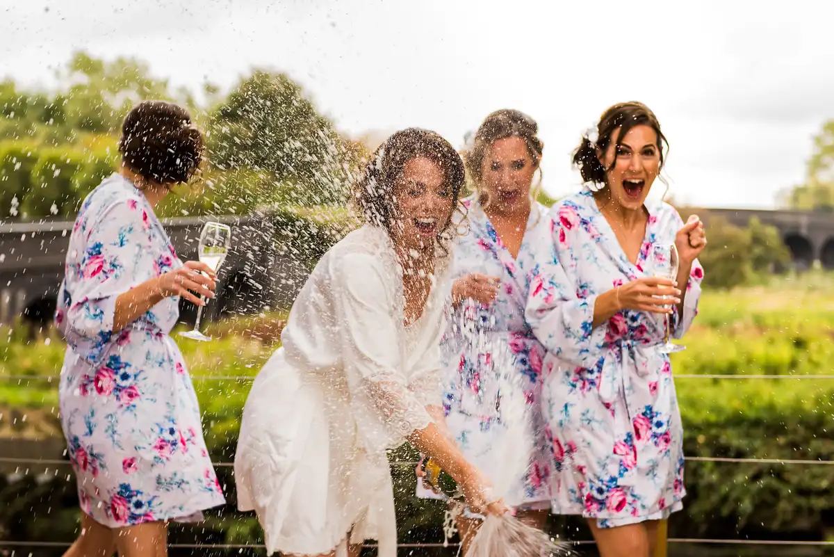 Bride and bridesmaids in floral robes shriek as champagne sprays over them in a garden.