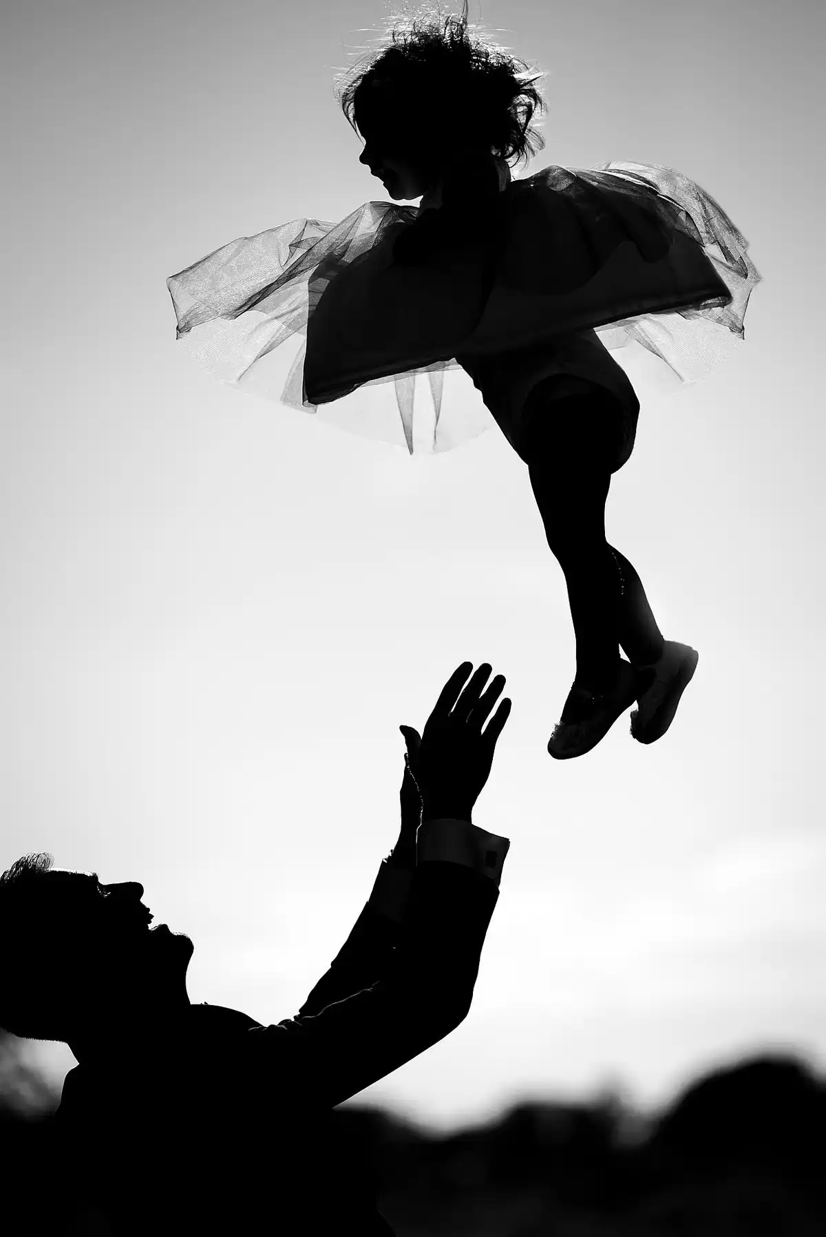 Black and white silhouette of a little girl in a tutu thrown high into the sky, a man's hands reaching up below.