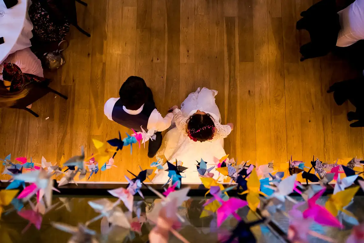 Seen from above, a page boy and a flower girl hold hands on a wooden floor beside a row of colourful paper cranes.