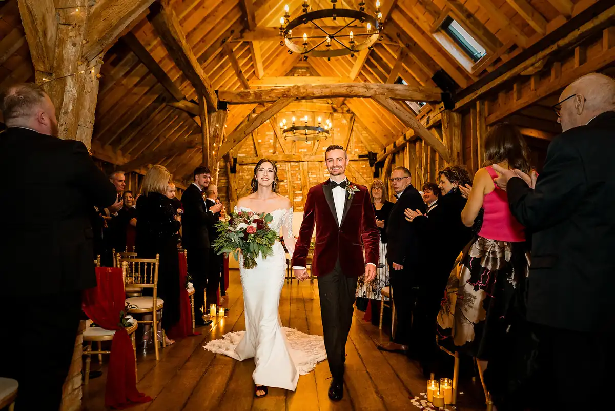 Bride and groom in a burgundy velvet jacket walk back down the aisle of a timber barn as guests clap.
