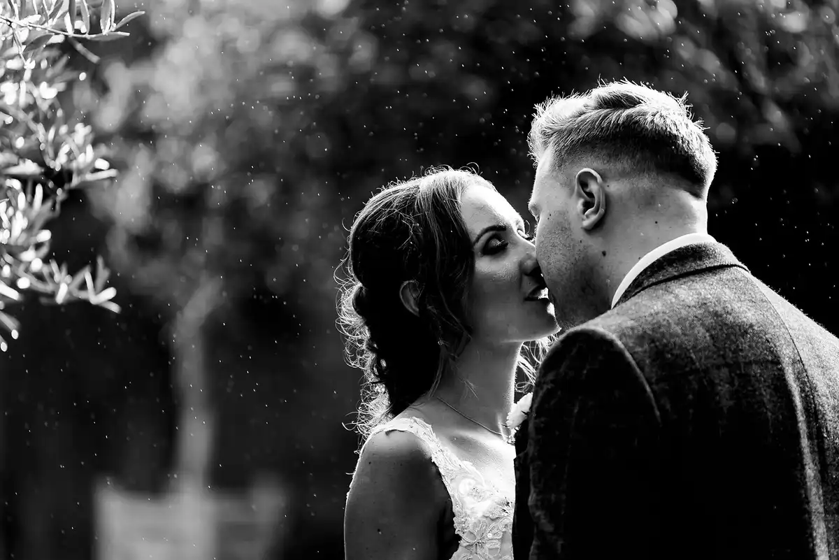 Black and white of the couple about to kiss, eyes closed, specks of rain catching the light behind them.