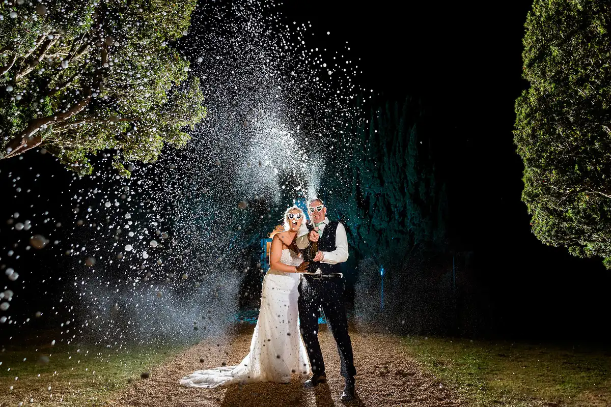 Couple in heart sunglasses spraying champagne high into the dark between two trees at night.