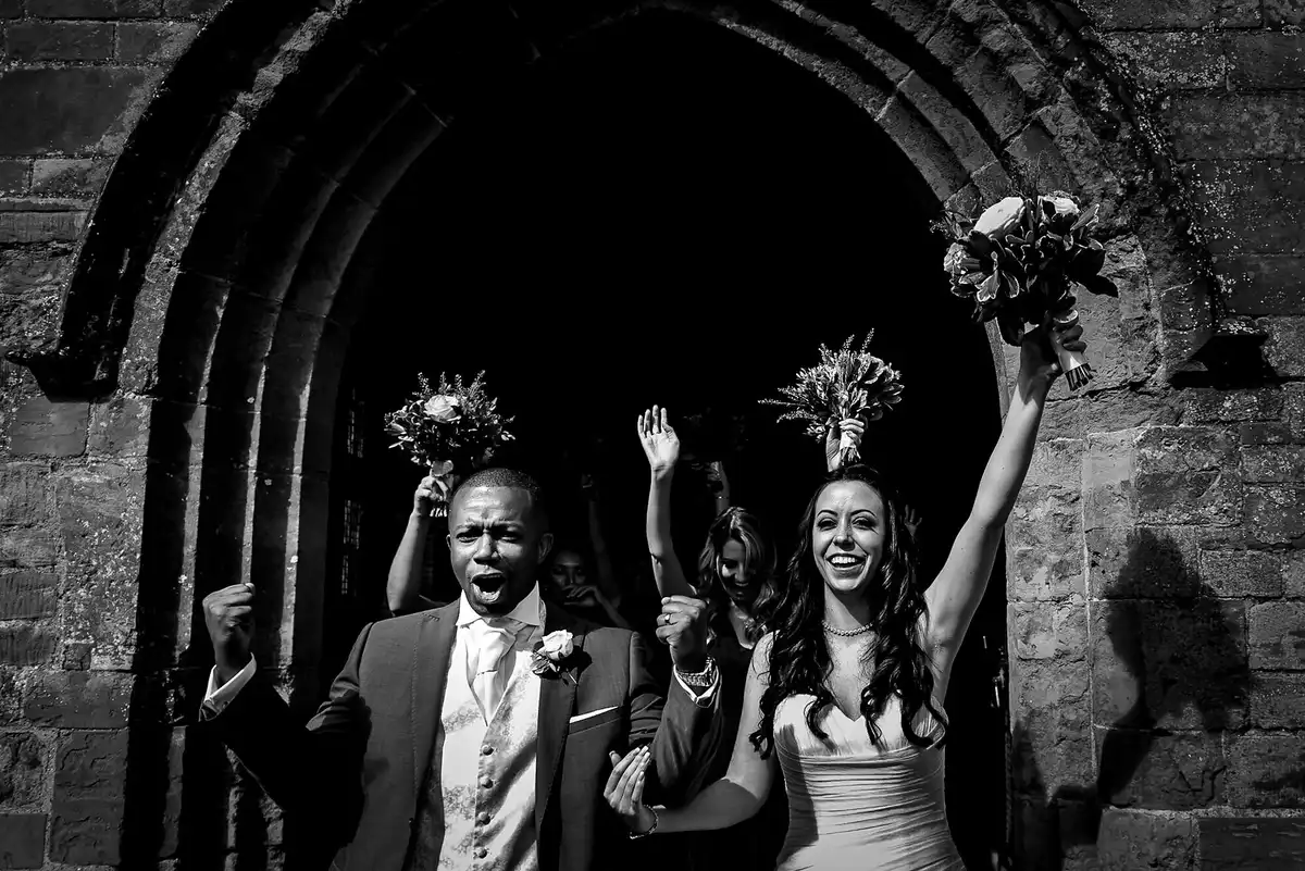 Black and white of the bride and groom bursting out of an arched church door, arms up, bridesmaids raising bouquets behind.