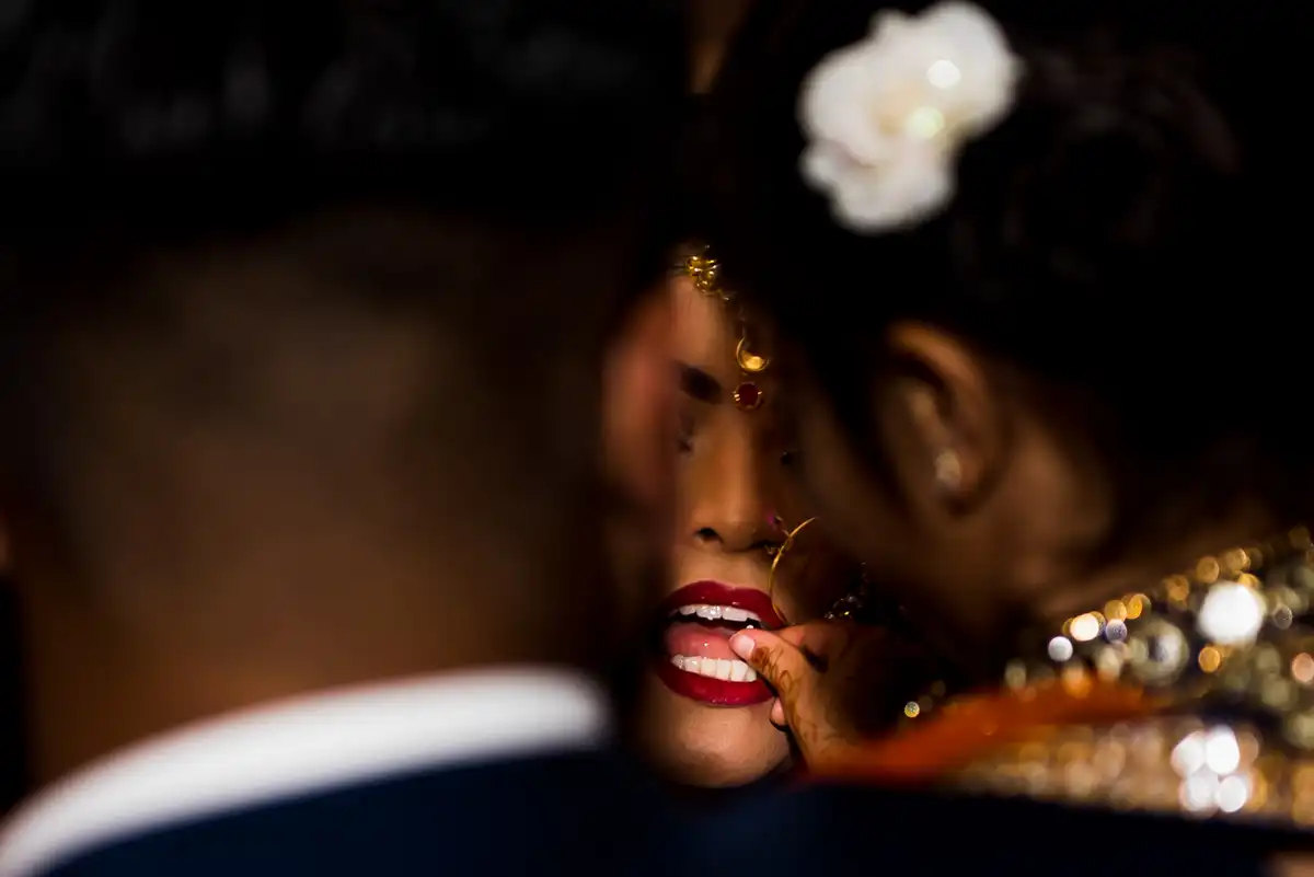 Close, intimate frame of a bride with red lips and gold jewellery leaning into the groom, his shoulder blurred in front.