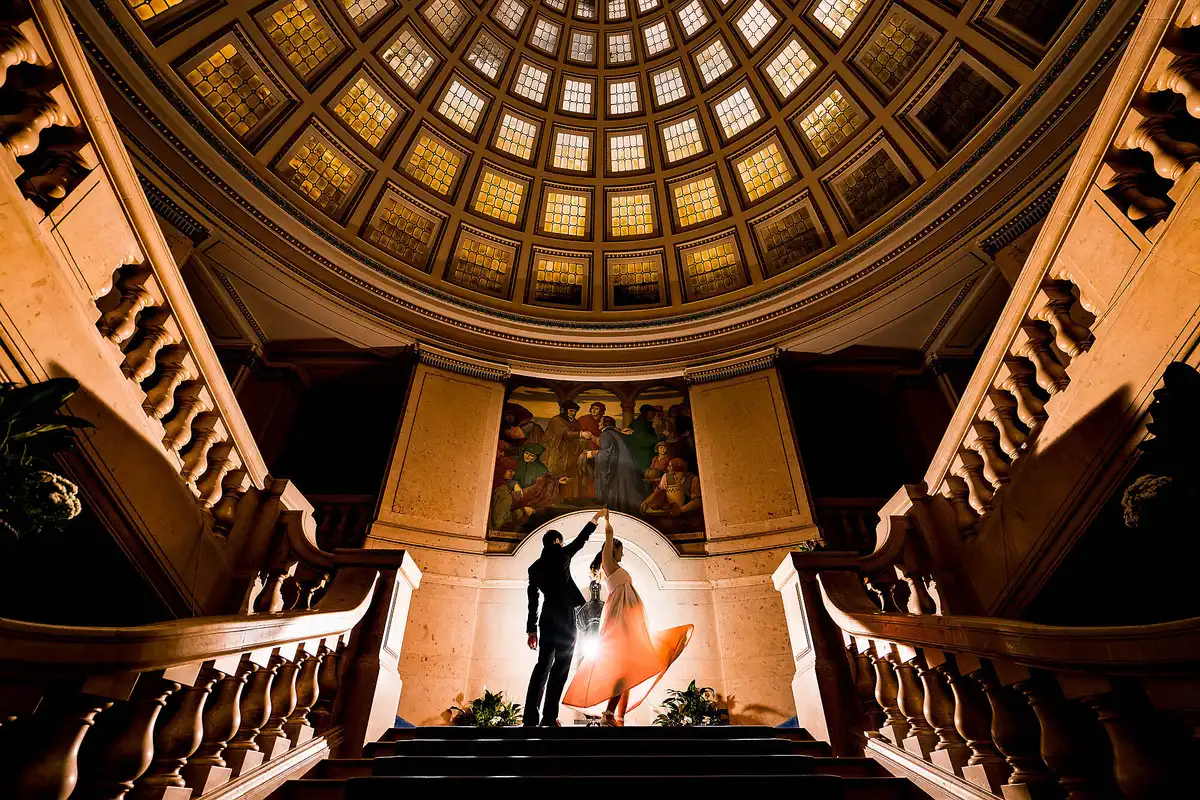 Couple dance at the top of a grand staircase beneath a glowing coffered dome, her skirt swirling orange.