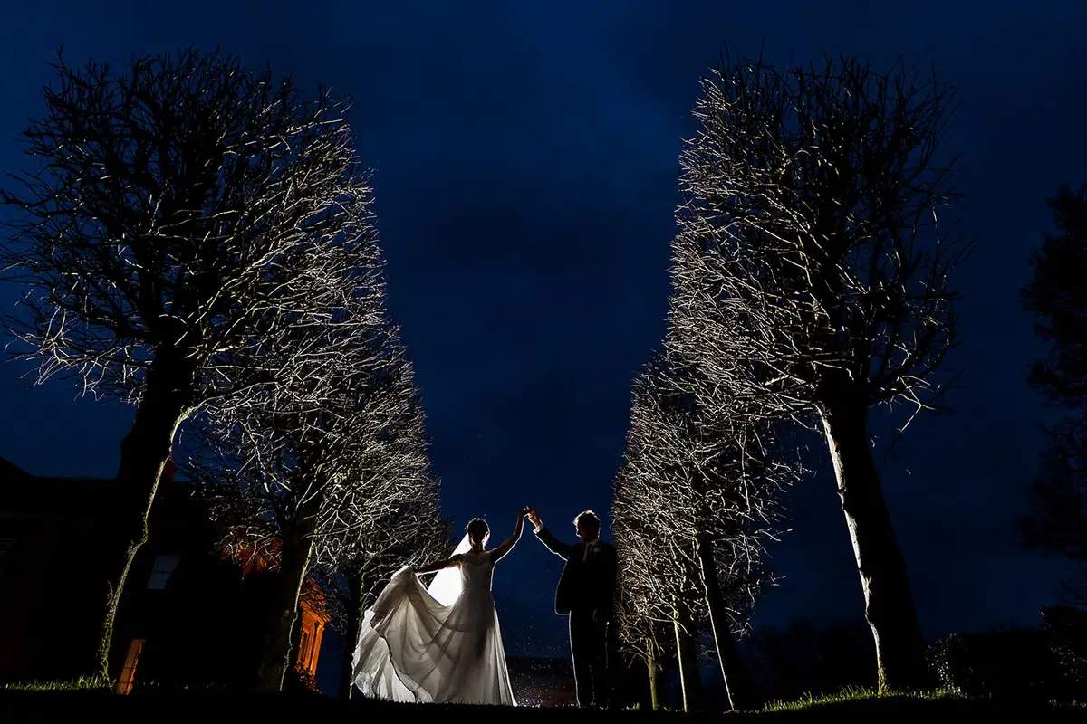 Bride and groom hold hands overhead between two rows of lit bare trees under a deep blue night sky.