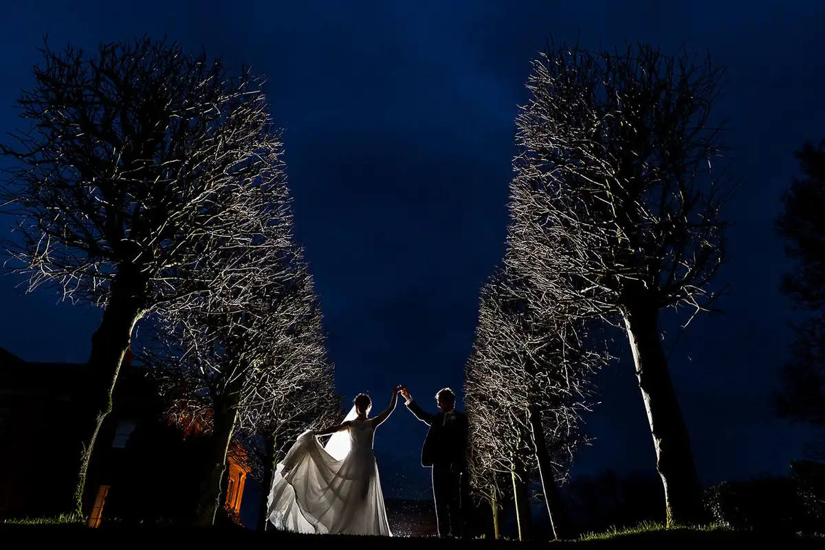 Bride and groom hold hands overhead between two rows of lit bare trees under a deep blue night sky.