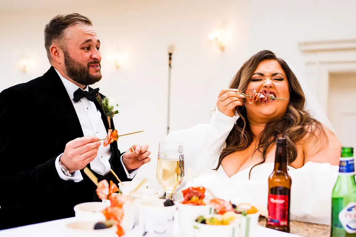 Bride and groom eating grazing skewers at the top table, the bride screwing up her face mid-bite.