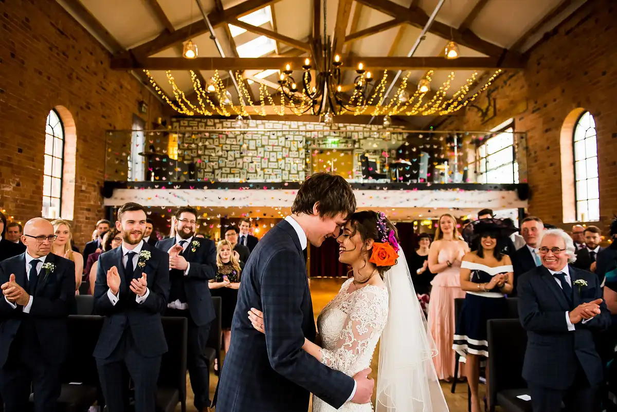 Bride and groom touch foreheads at the front of a red brick hall strung with fairy lights as guests stand and clap.