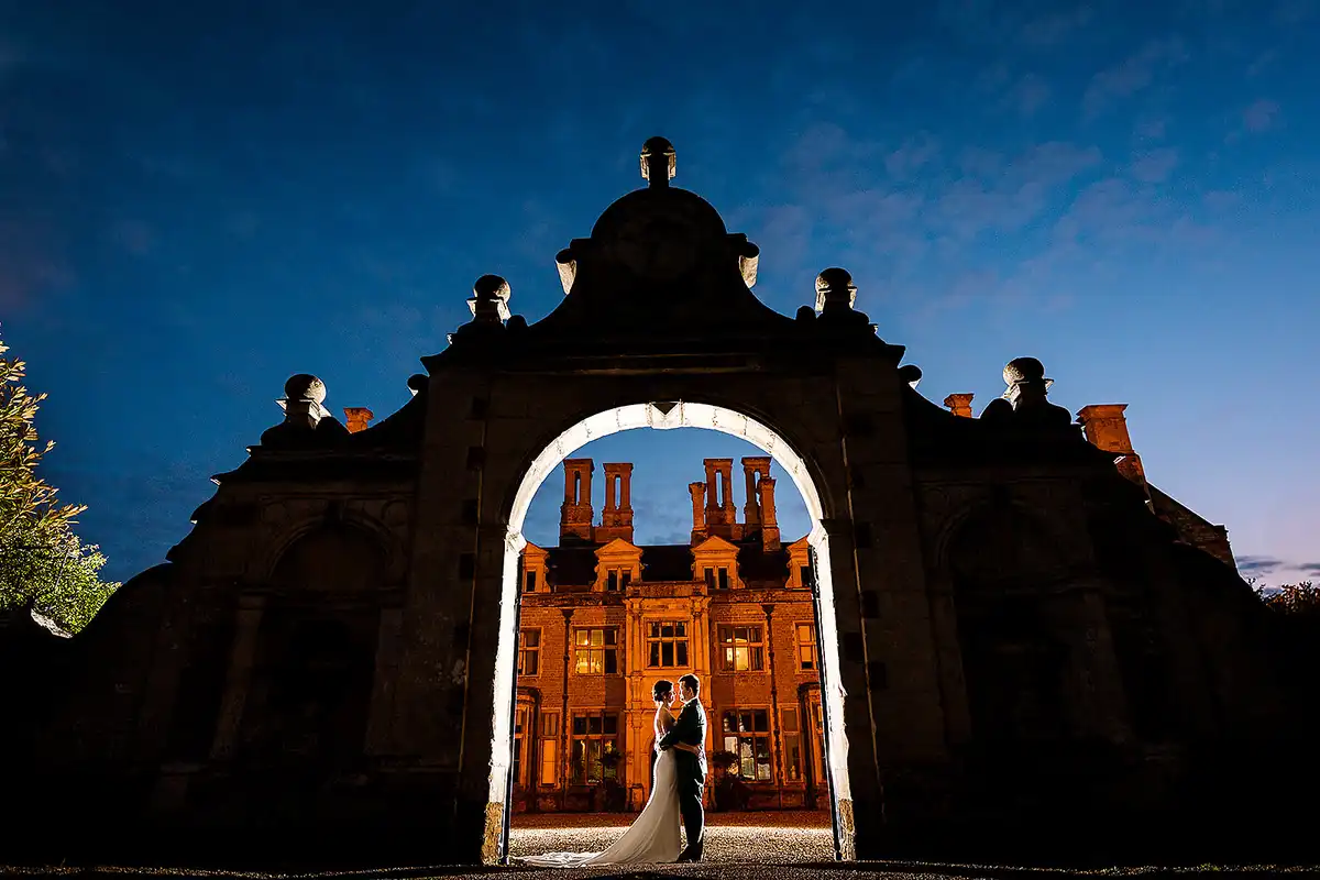 Couple framed by a lit stone gatehouse arch at dusk, a red brick manor glowing behind them.