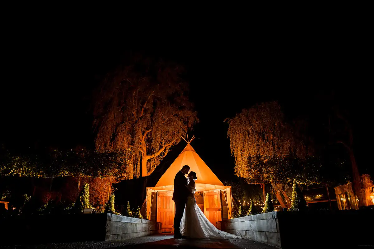Couple embrace in silhouette in front of a glowing tipi and lit willow trees at night.