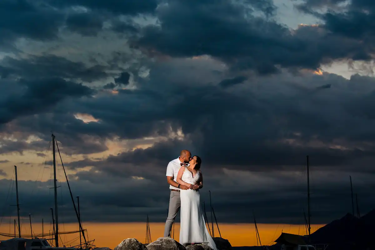 Couple kissing on rocks at a harbour under heavy storm clouds, yacht masts and an orange sunset behind.