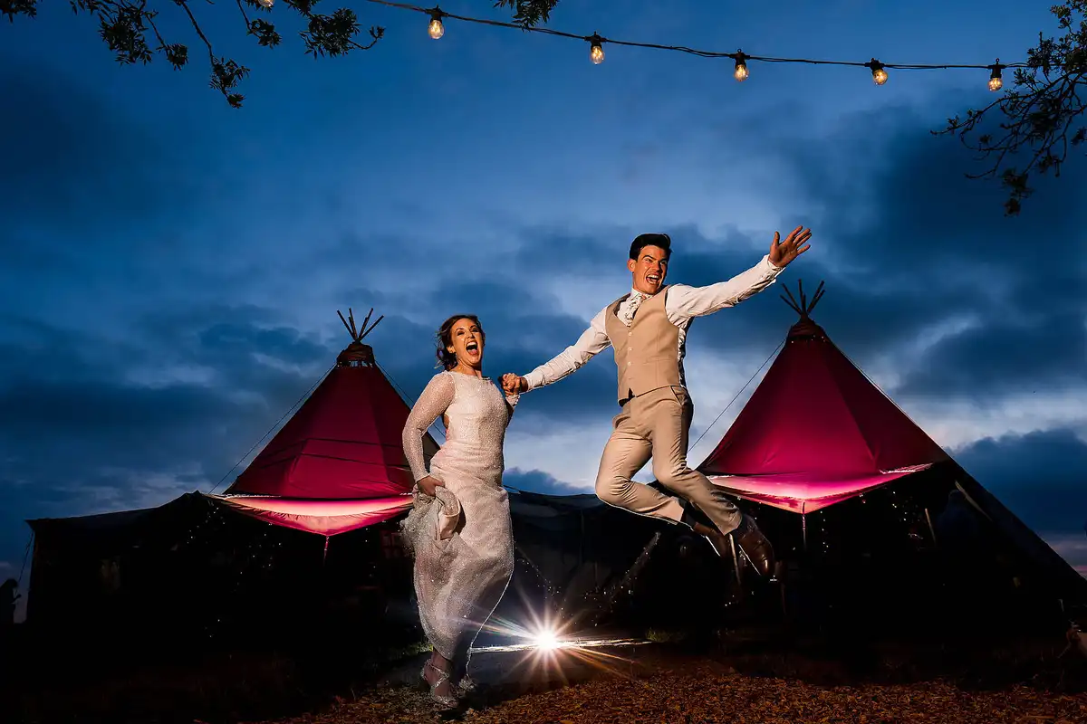 Groom leaps high and the bride shrieks between two pink-lit tipis under a stormy dusk sky.
