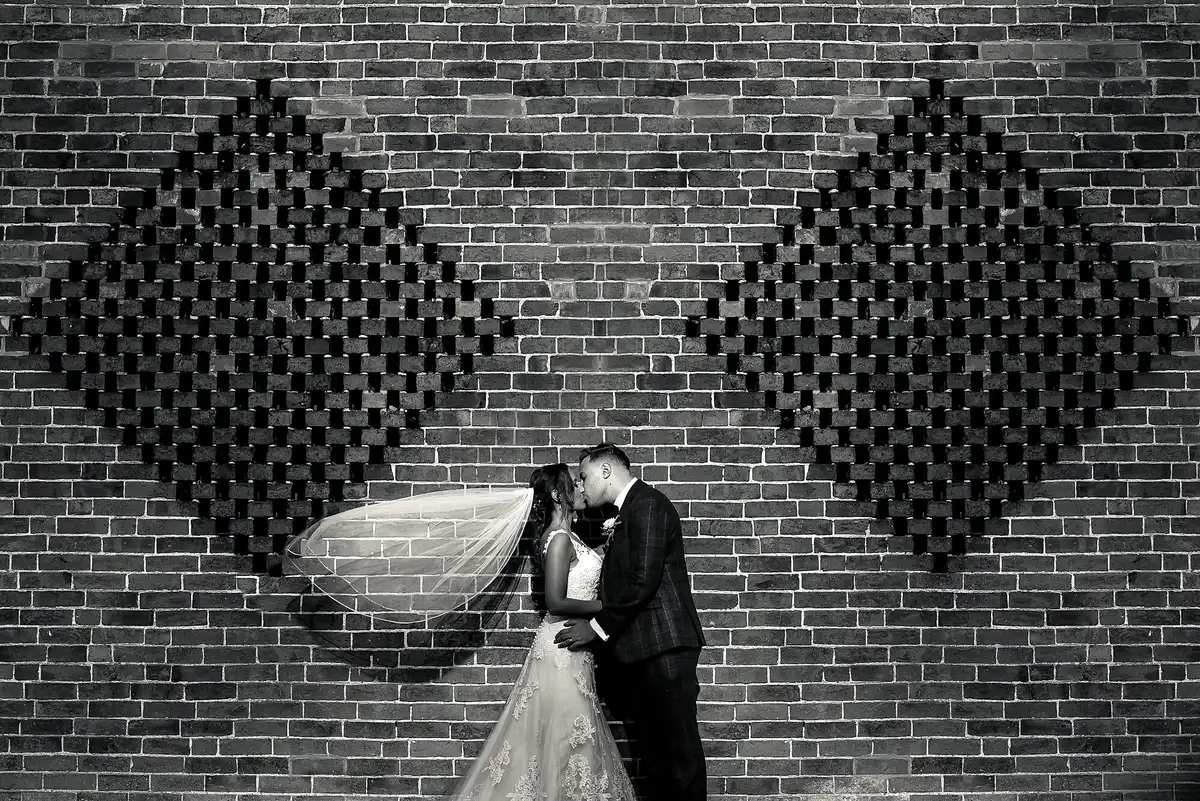 Black and white of the couple kissing against a patterned brick wall as the bride's veil blows out sideways.