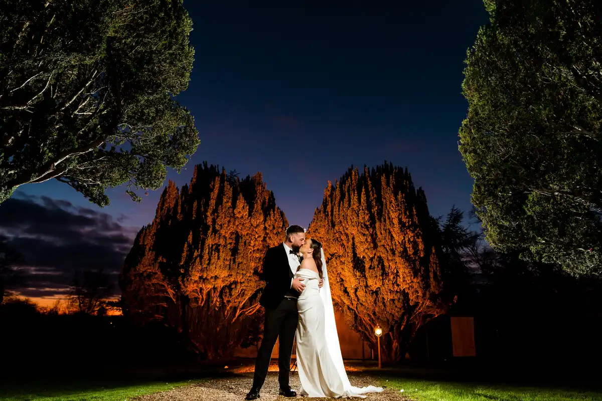 Couple kissing at dusk in front of two yew trees lit orange, a blue sky fading behind them.
