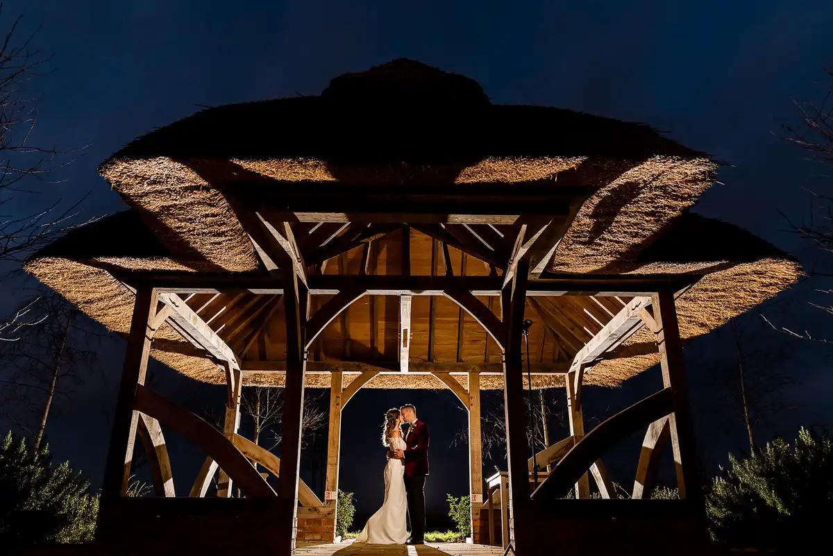 Low view of the couple kissing under a lit thatched timber gazebo against a dark blue night sky.