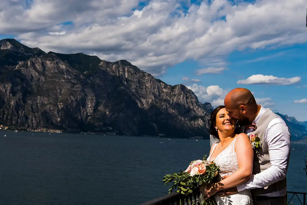 Groom kisses the laughing bride's head on a terrace above a lake, big mountains and clouds behind.