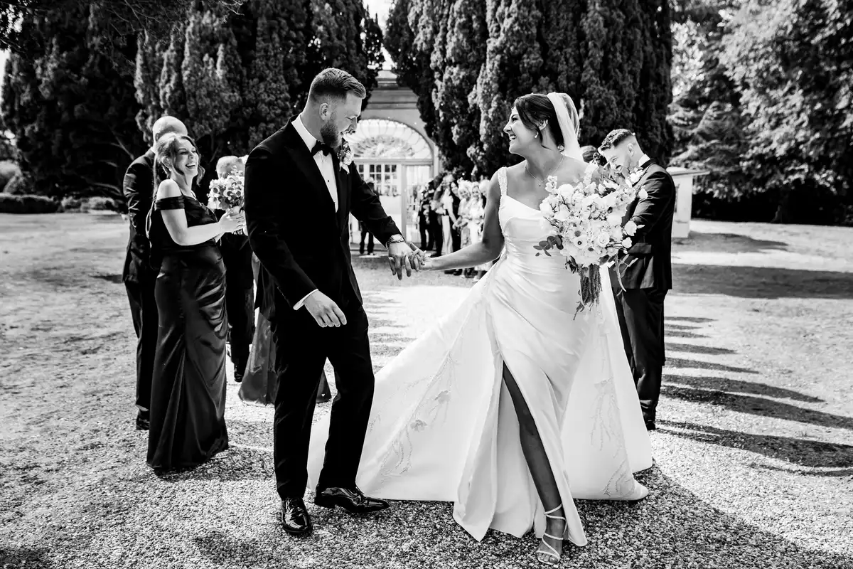 Black and white of the bride and groom laughing hand in hand on a gravel drive, wedding party behind.