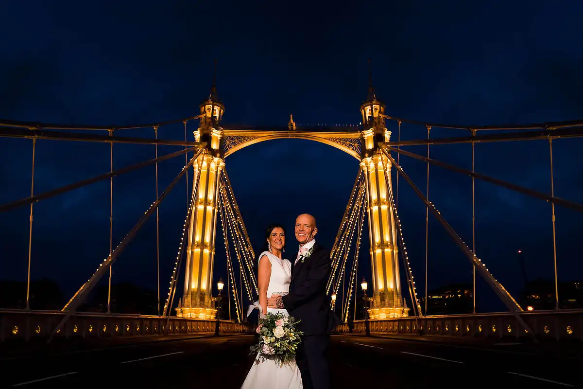 Couple stand on a suspension bridge at night, its golden lit towers and cables framing them against a blue sky.