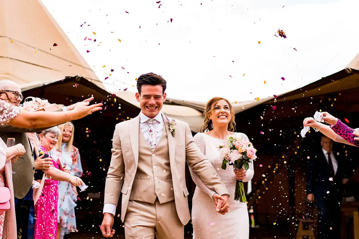 Groom in a cream suit and the bride grin as guests throw petal confetti outside a tipi.