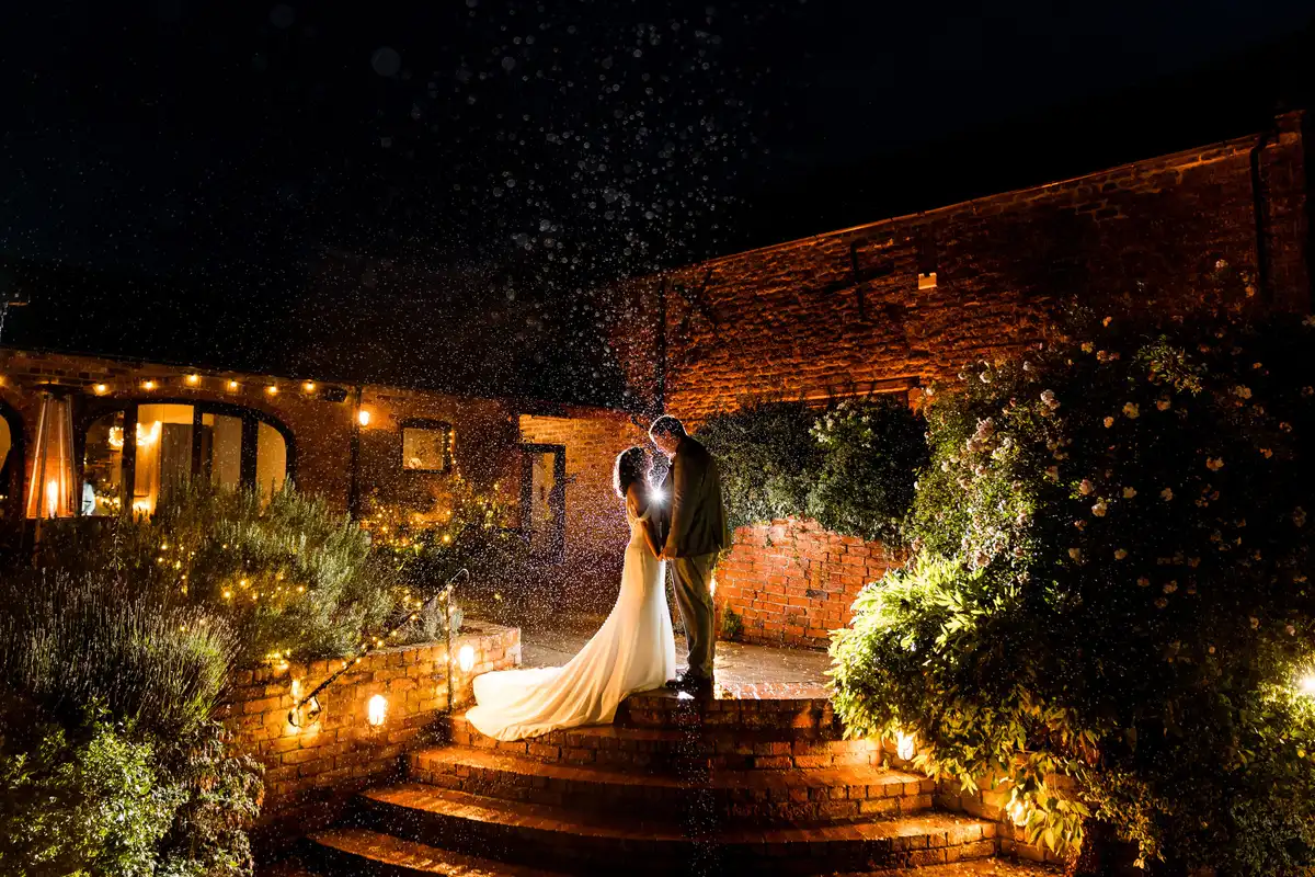 Couple face each other on brick steps at night as rain falls, fairy lights along the walls.