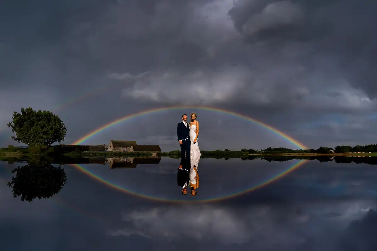 Bride and groom stand under a full rainbow and storm clouds, the whole scene mirrored in still water.