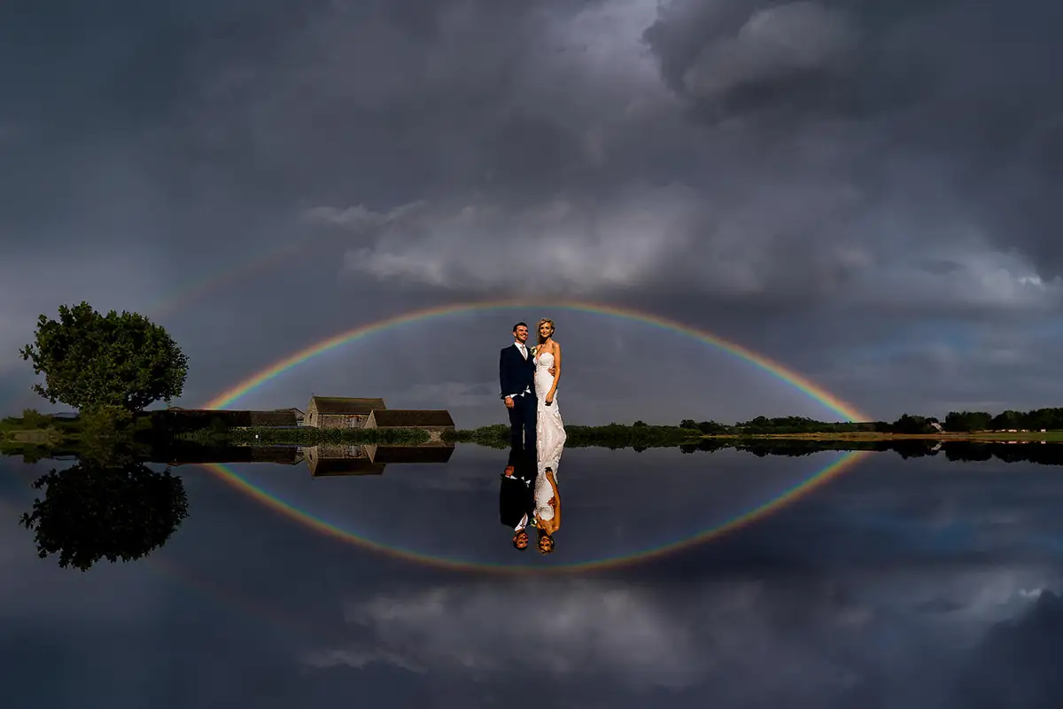 Bride and groom stand under a full rainbow and storm clouds, the whole scene mirrored in still water.