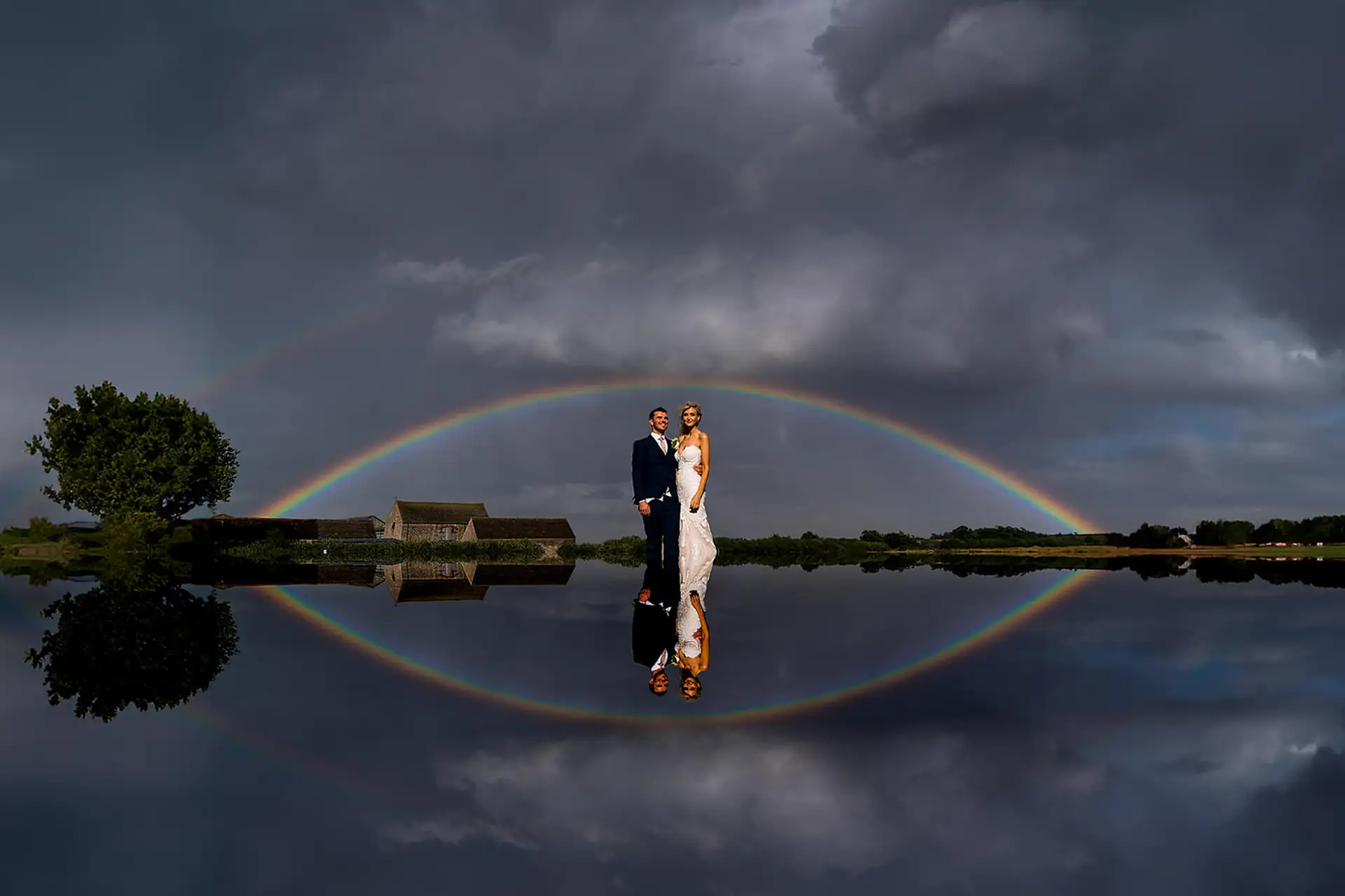 Bride and groom stand under a full rainbow and storm clouds, the whole scene mirrored in still water.