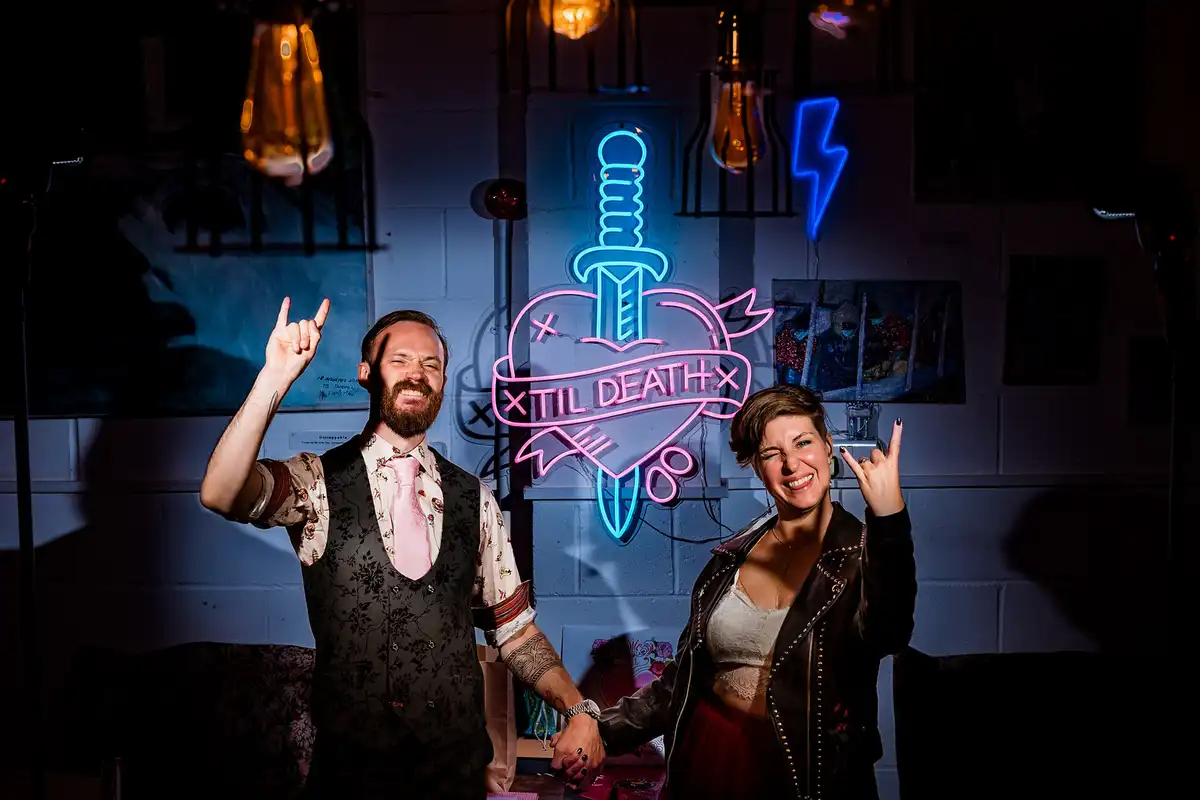 Couple holding hands and throwing rock-on hand signs beside a neon heart sign reading Til Death.