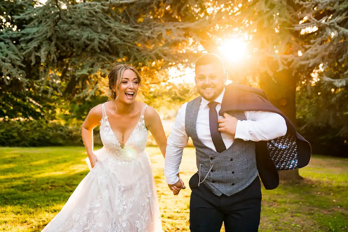 Bride and groom laugh as they run hand in hand across a lawn, sun bursting through the trees, his jacket over his shoulder.