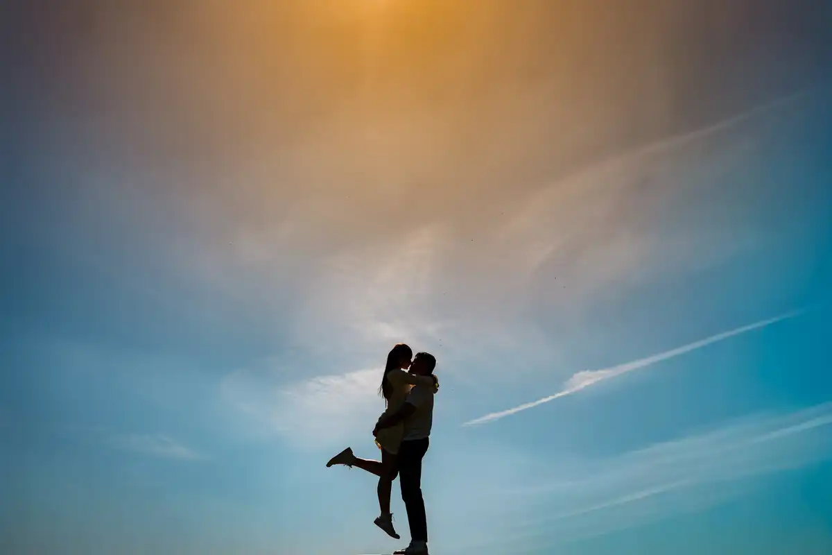 Couple in silhouette against a blue and gold sky, one lifted up and kissing the other.
