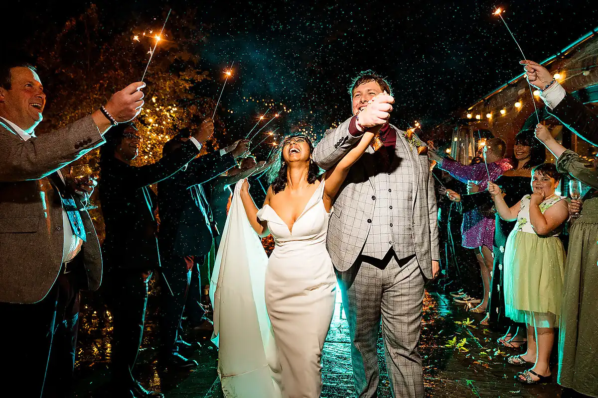 Bride and groom raise joined hands through a tunnel of sparklers at night, guests cheering either side.