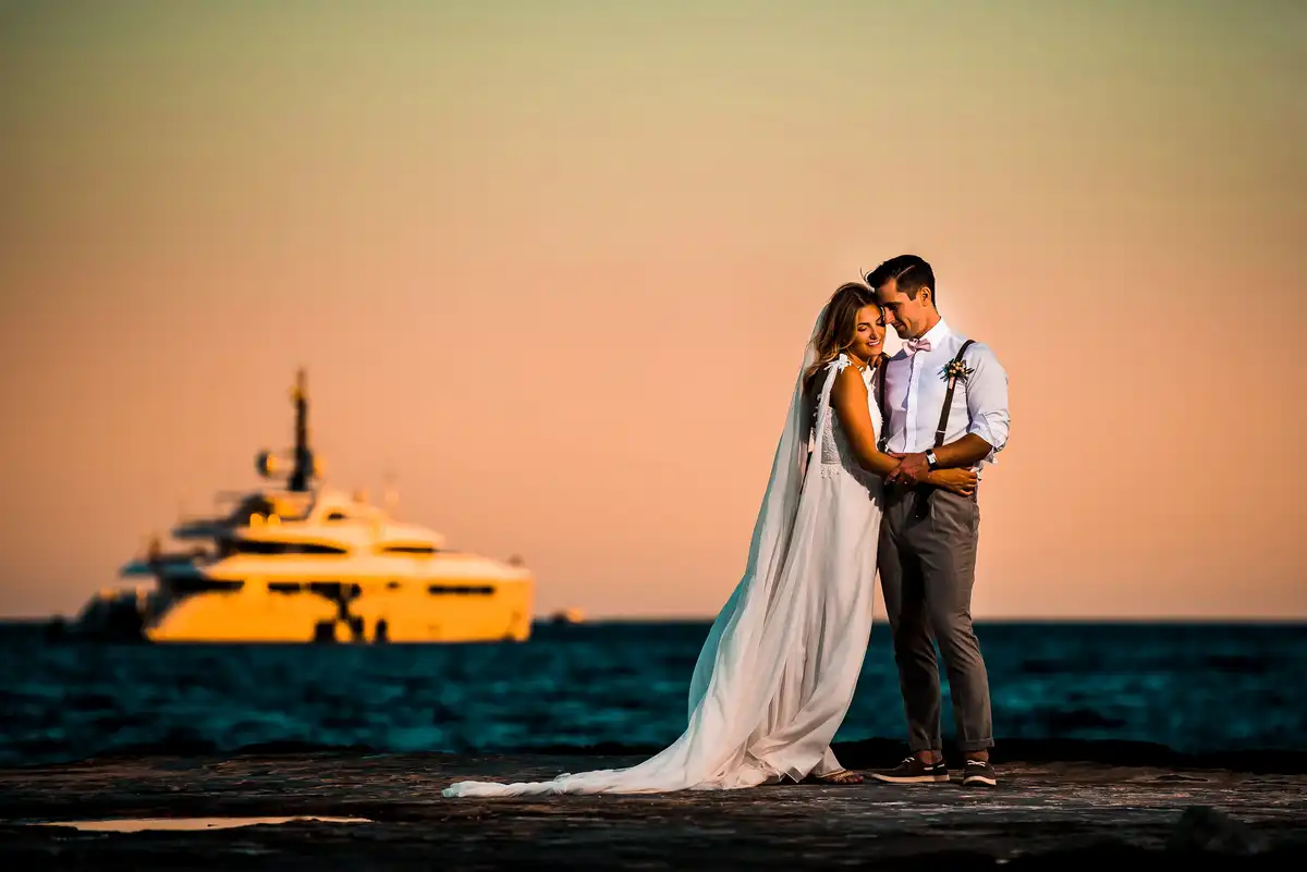 Couple cuddle on a rocky shore at sunset, a big yacht glowing orange on the sea behind them.