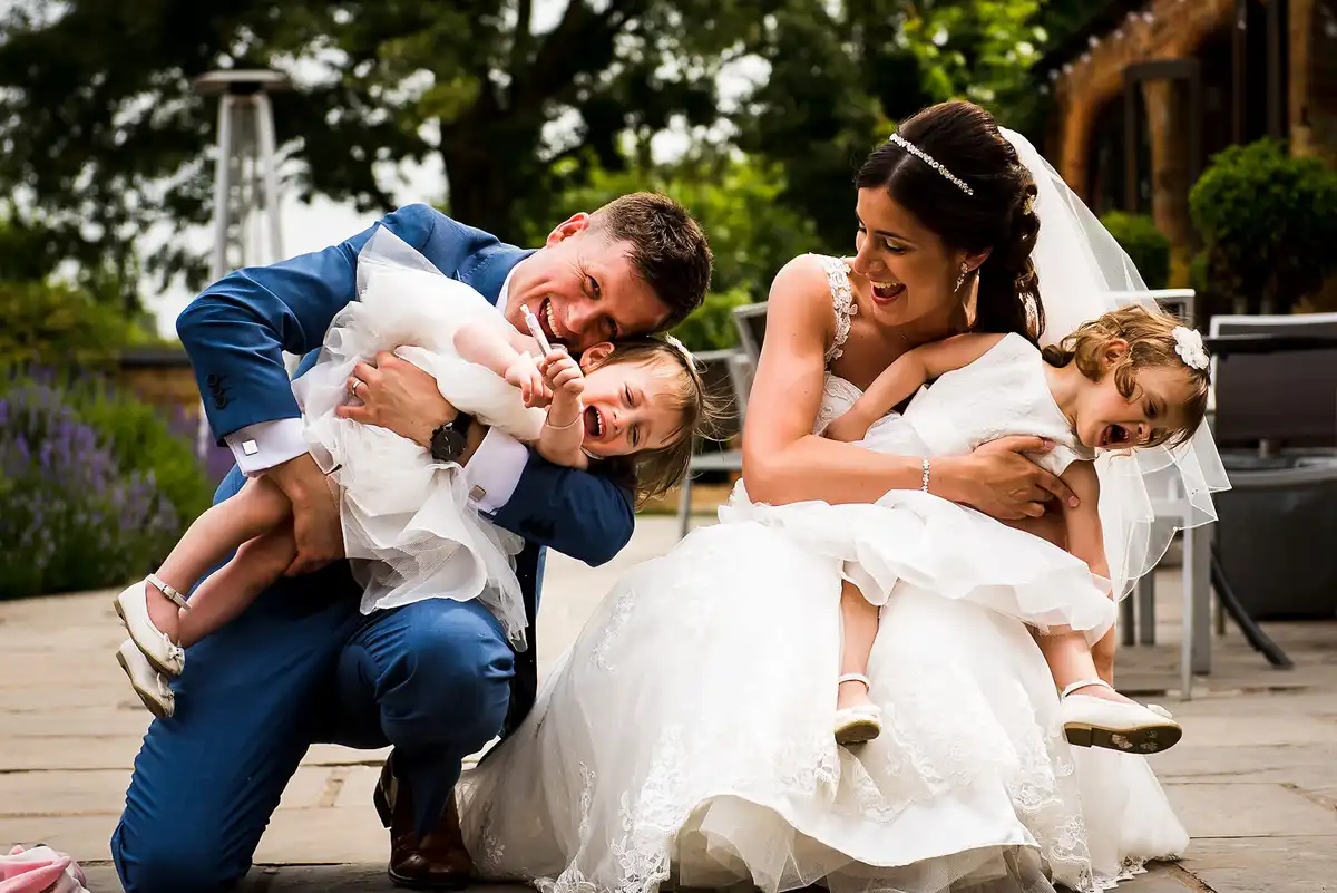 Bride and groom crouch and tickle two shrieking flower girls on a garden terrace.