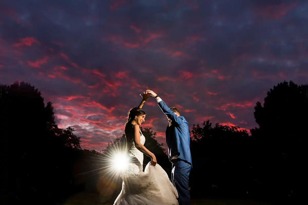 Groom twirls the bride under a red streaked sunset sky, a light flaring behind her dress.