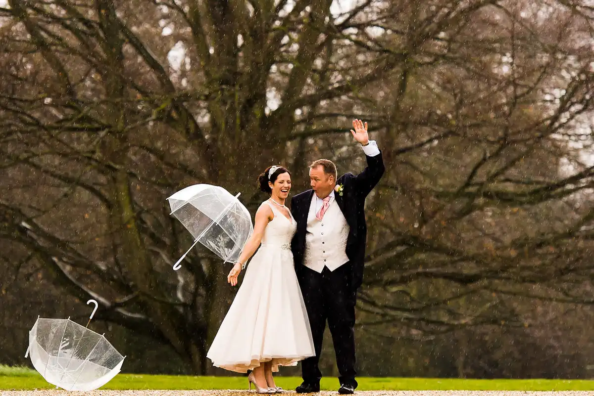 Bride laughing with a clear umbrella as the groom waves, a second umbrella blown on to the grass, bare trees behind.