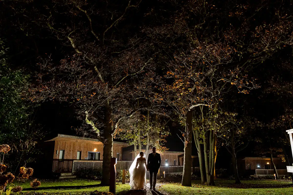 Bride and groom walk hand in hand along a path under big autumn trees at night, timber lodges behind.