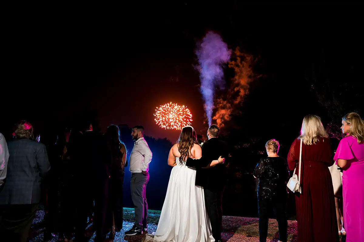 Bride and groom seen from behind, arms around each other, watching fireworks with guests at night.