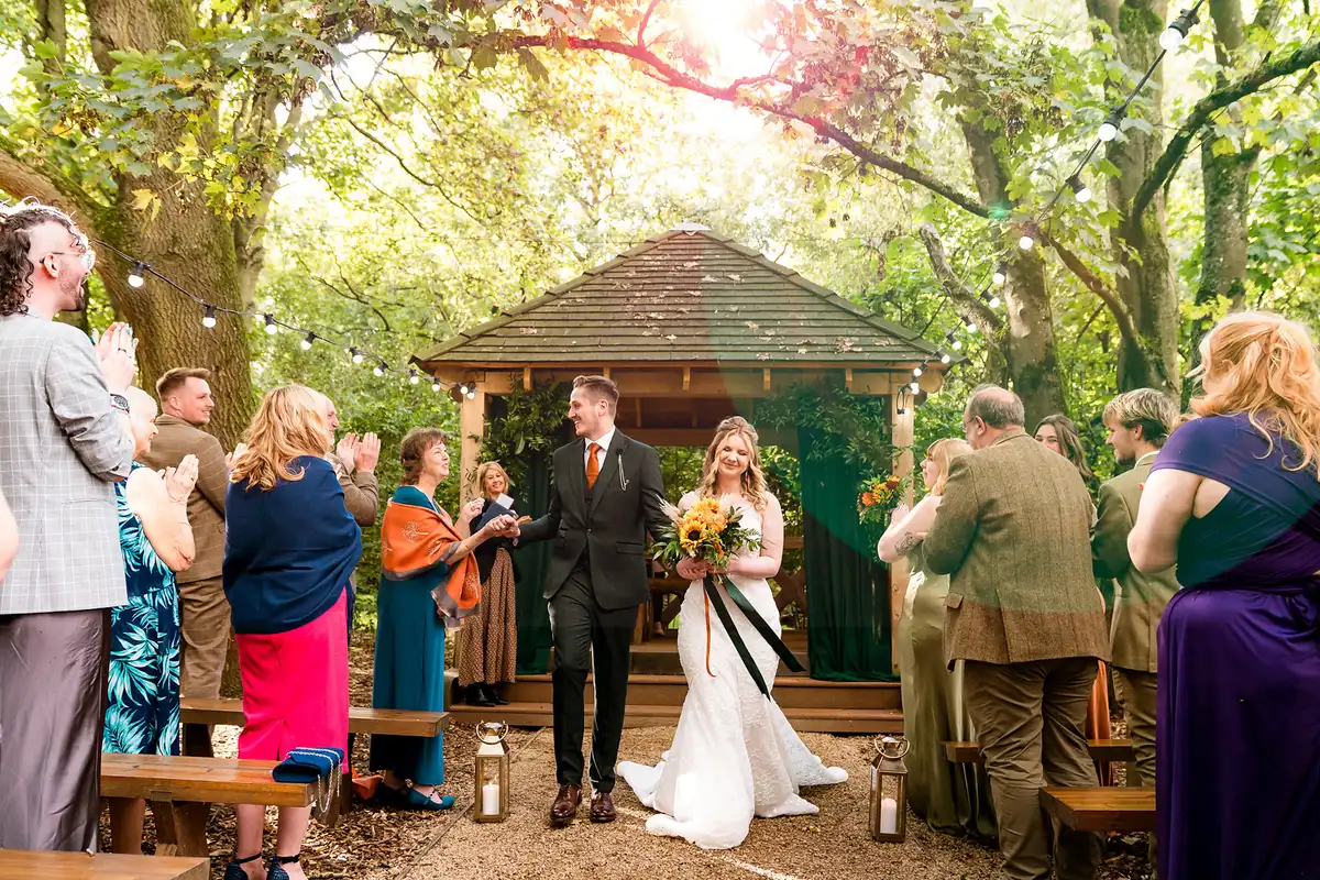 Newlyweds walk back down a woodland aisle from a wooden gazebo as guests clap in soft sunlight.