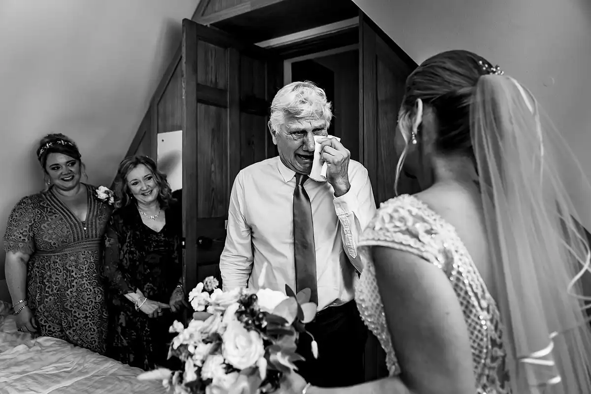 Black and white of an older man wiping away tears as he sees the bride in her dress, two women smiling.