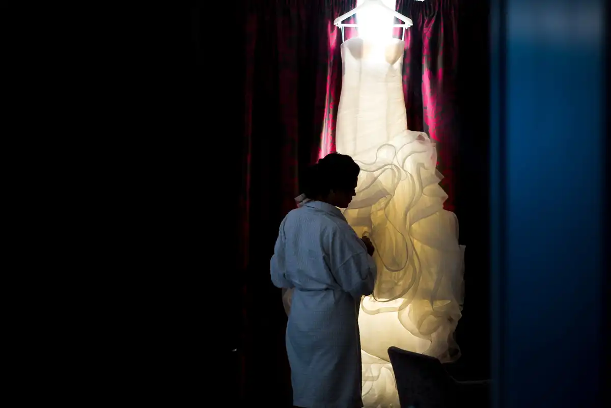 Woman in a dressing gown looks up at a wedding dress hanging in a bright window in a dark room.