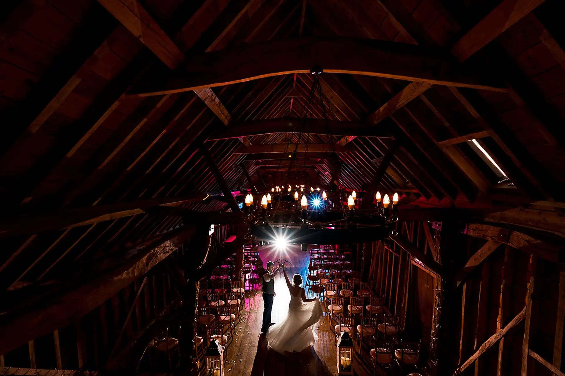 First dance seen from the rafters of a dark timber barn, the couple backlit beneath a candle chandelier.