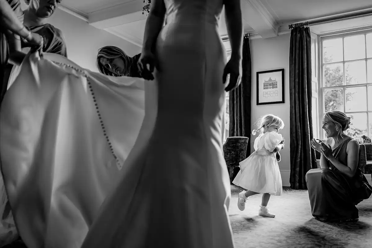 Black and white of bridesmaids buttoning the bride's dress while a little flower girl dances past.