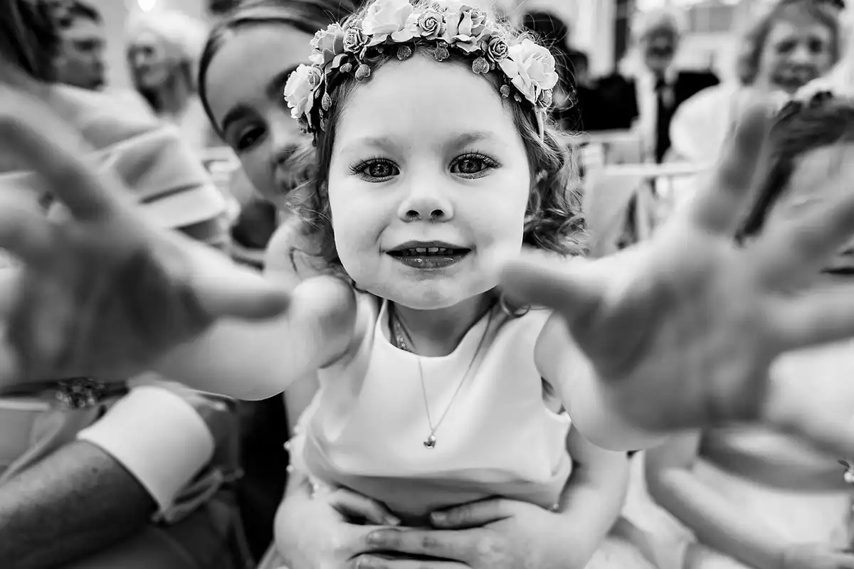 Black and white close-up of a flower girl in a rose crown reaching both hands towards the camera.
