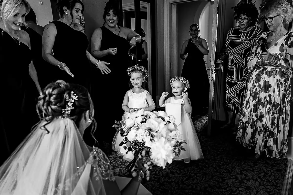 Black and white of two flower girls carrying a big bouquet towards the bride as women watch from the doorway.
