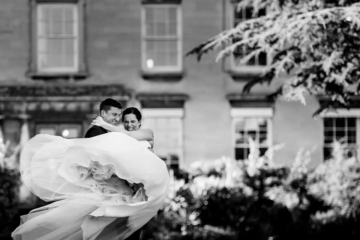 Black and white of the groom carrying the laughing bride, her full skirt billowing, in front of a manor house.